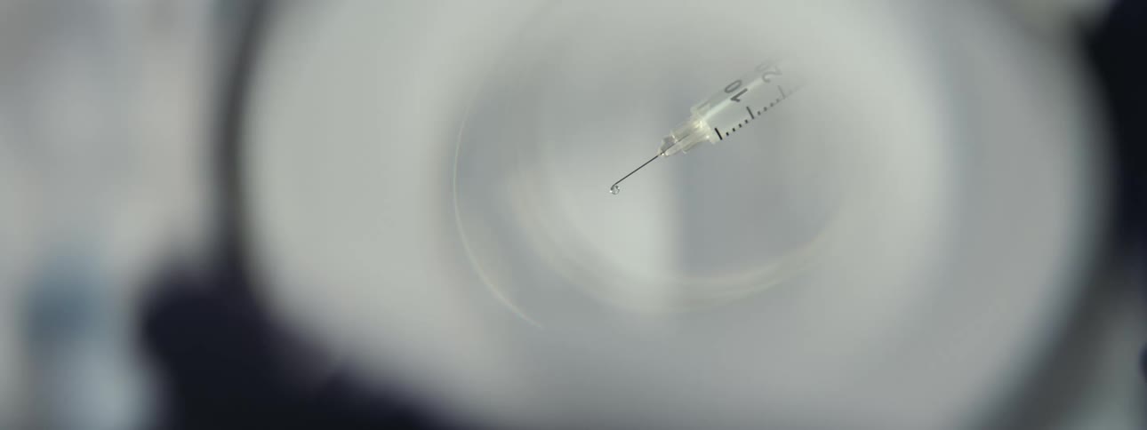 An artistic close-up of a syringe needle with a single droplet of medicine seen through a distorted lens