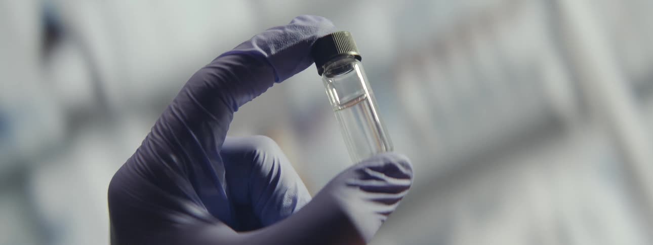 A scientist in a sterile lab wearing purple gloves carefully examines a small vial containing a clear liquid sample