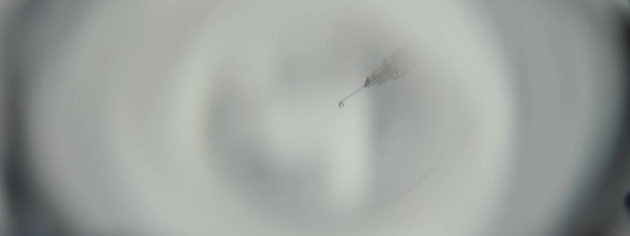 A close-up of a syringe with a droplet of medicine on the needle's tip, viewed through a magnifying lens