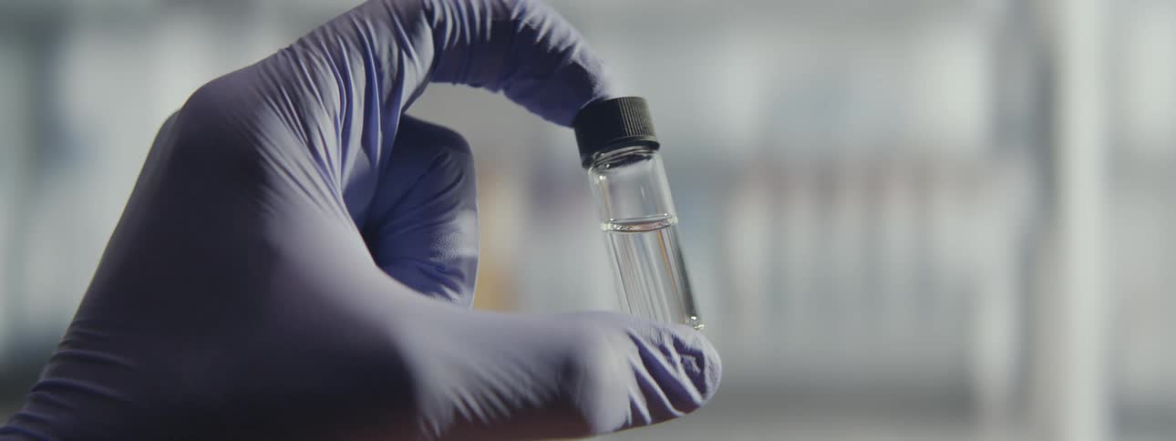 A scientist's gloved hand holds up a small glass vial filled with a clear liquid for inspection in a laboratory