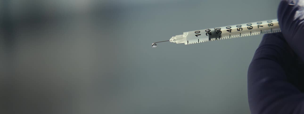 A close-up, slow-motion shot shows a single drop of clear liquid falling from the tip of a syringe needle held by a gloved hand