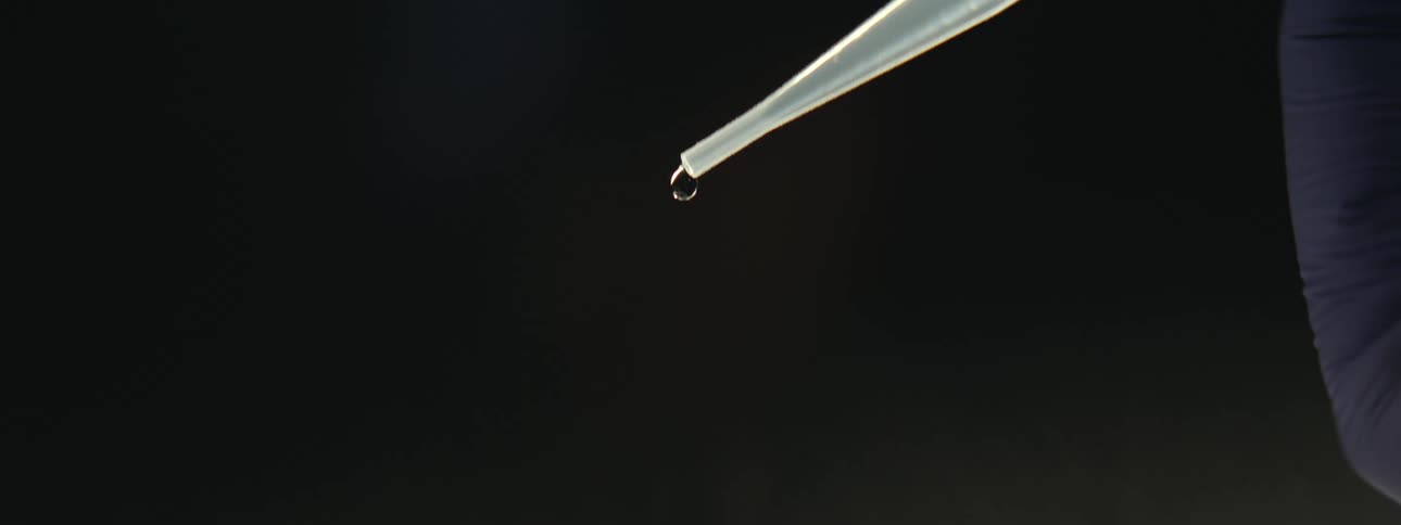 A single drop of clear liquid falls from a pipette in extreme slow motion against a black background