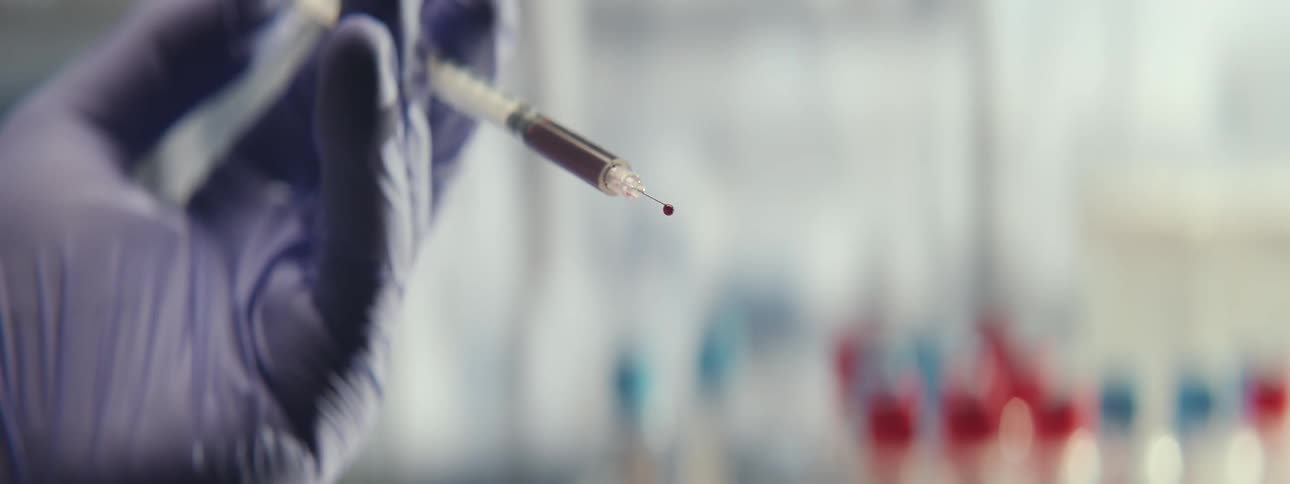 In slow motion, a drop of blood falls from a syringe held by a gloved hand in a sterile medical laboratory