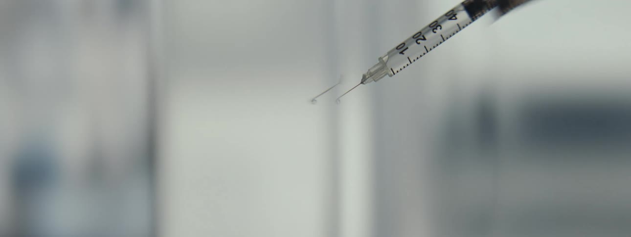 A single drop of a clear liquid hangs from the tip of a syringe needle in a macro close-up shot