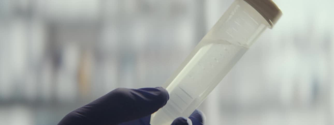A close-up shot of a gloved hand holding and swirling a liquid sample inside a plastic vial in a laboratory setting