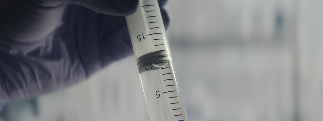 A close-up shot shows a gloved medical professional slowly emptying a syringe filled with clear liquid