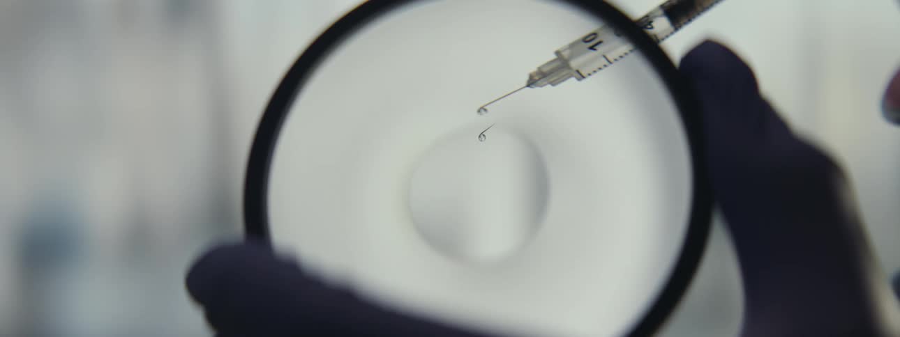 A gloved hand holds a magnifying glass over a syringe needle with a single drop of liquid on its tip