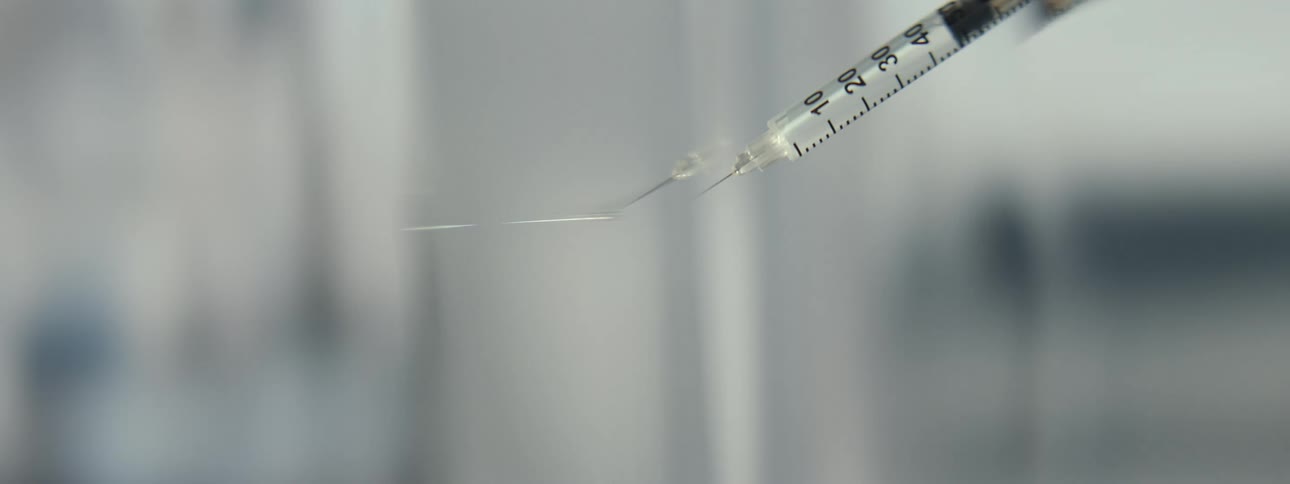A slow motion macro shot shows a clear liquid forming a droplet on the tip of a syringe needle in a sterile environment