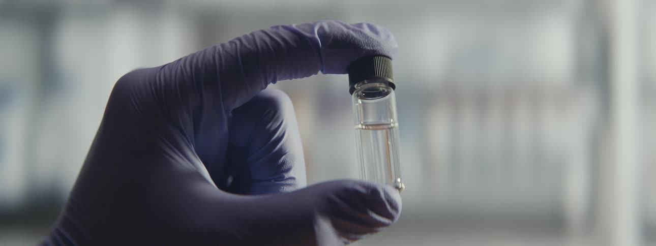 Close-up of a scientist's gloved hand holding and inspecting a small glass vial with a clear liquid sample in a laboratory