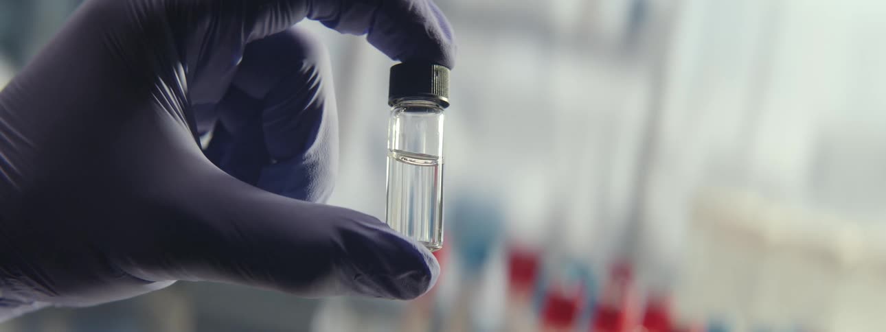 A scientist in a sterile lab holds up a small glass vial containing a clear liquid sample for inspection