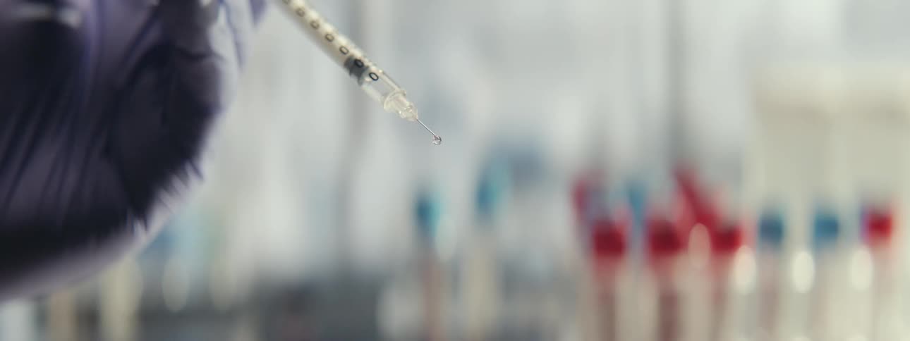 A gloved hand holds a syringe as a single drop of clear liquid forms on the needle and falls in slow motion in a laboratory