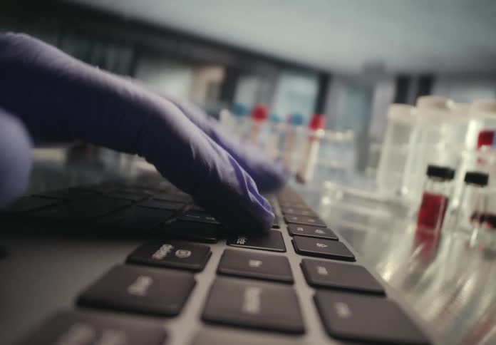 A stylistic low-angle shot of a researcher's gloved hands typing on a laptop keyboard in a scientific laboratory