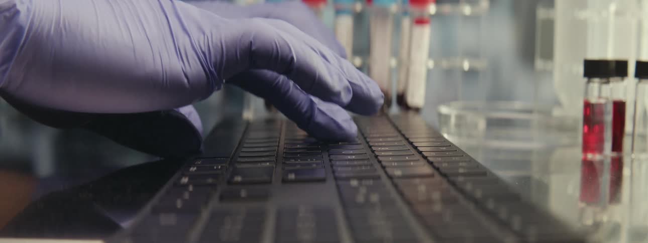 A close-up of a scientist's gloved hands typing on a laptop keyboard in a laboratory with blood samples in the background