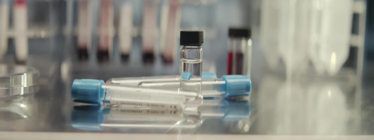 Medical test tubes and a vial containing clear liquid sit on a counter in a laboratory, with blood samples in the background