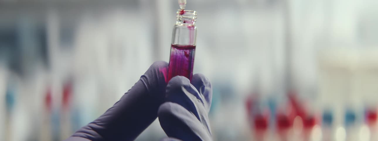 Close-up of a scientist in a lab adding red liquid from a pipette into a glass vial with purple solution, causing a chemical reaction