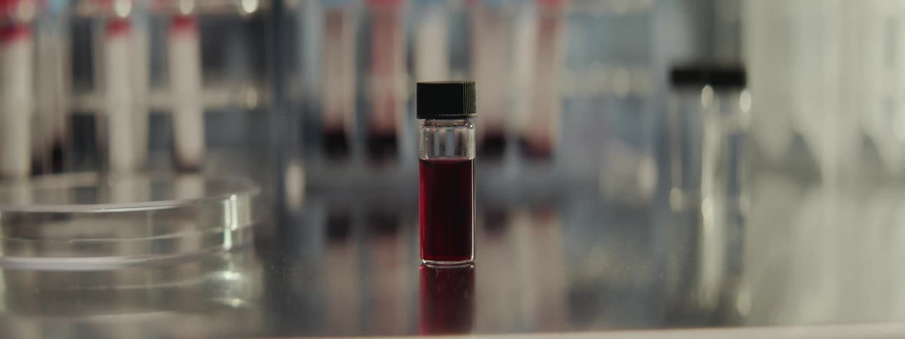 A slow dolly out reveals a close-up of a small glass vial of blood on a metal countertop in a sterile research laboratory