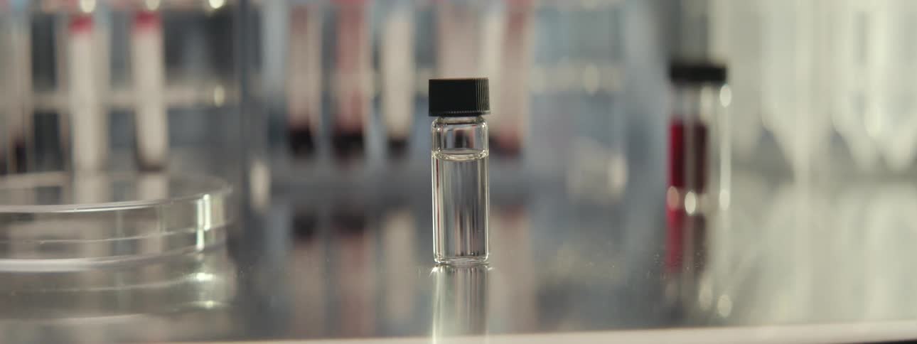A close-up of a small glass vial with a clear liquid sits on a sterile surface in a science laboratory