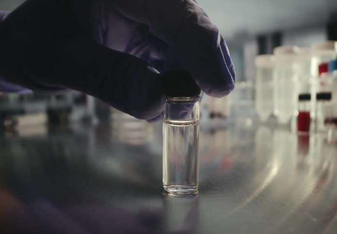 A gloved hand carefully picks up a small glass vial of clear liquid from a metal table in a sterile lab environment