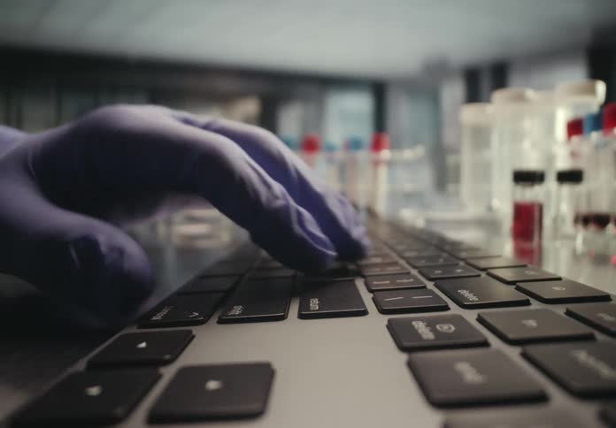 A close-up of a gloved hand typing on a keyboard in a scientific research laboratory with vials and test tubes in the background