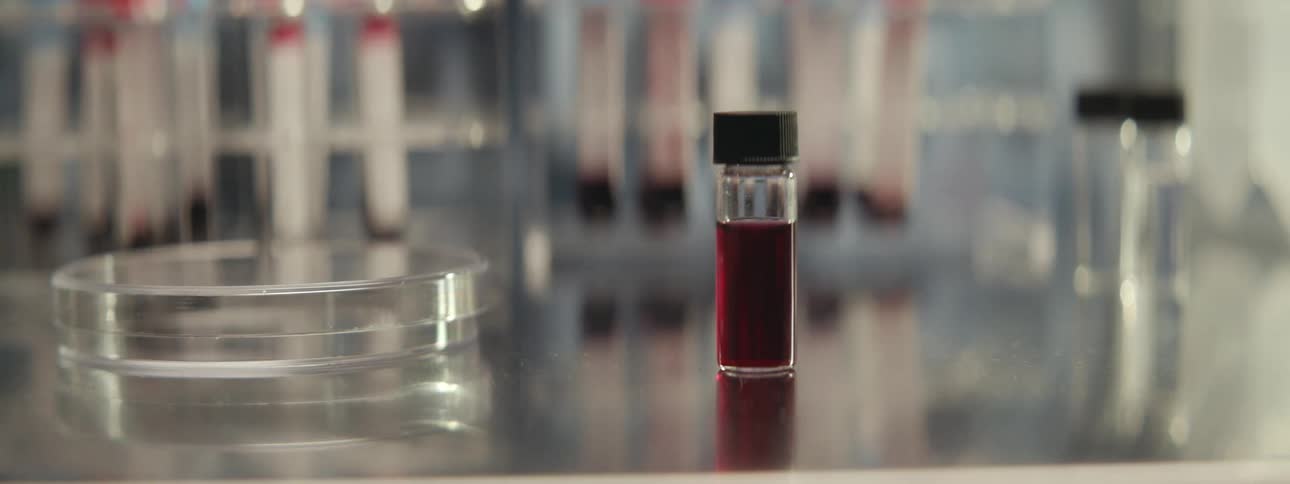 A close-up pan in a laboratory reveals racks of test tubes with blood samples, and a single vial on a metal countertop
