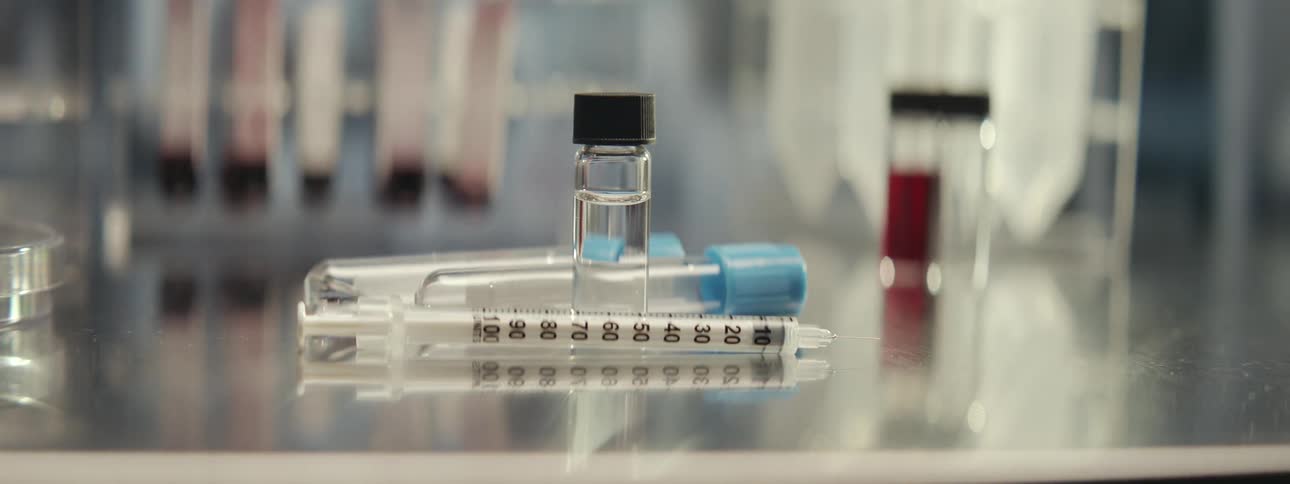 A slow focus pull reveals a syringe, vial, and test tubes on a reflective surface in a medical laboratory