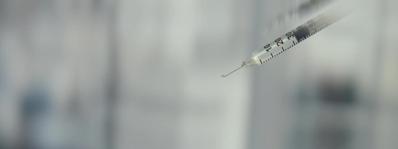 A close-up, slow-motion shot shows a liquid vaccine dripping from the tip of a syringe needle in a sterile environment