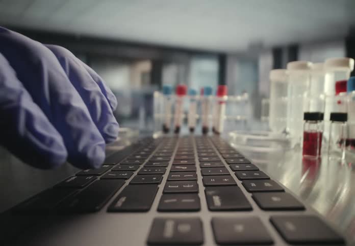 A dolly shot pulls away from a researcher's gloved hands typing on a laptop in a science lab with test tubes