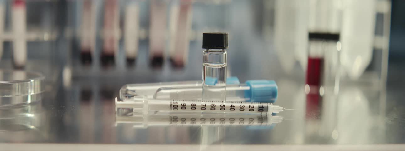 A sterile syringe, a vial of clear vaccine, and test tubes rest on a metal counter in a modern laboratory