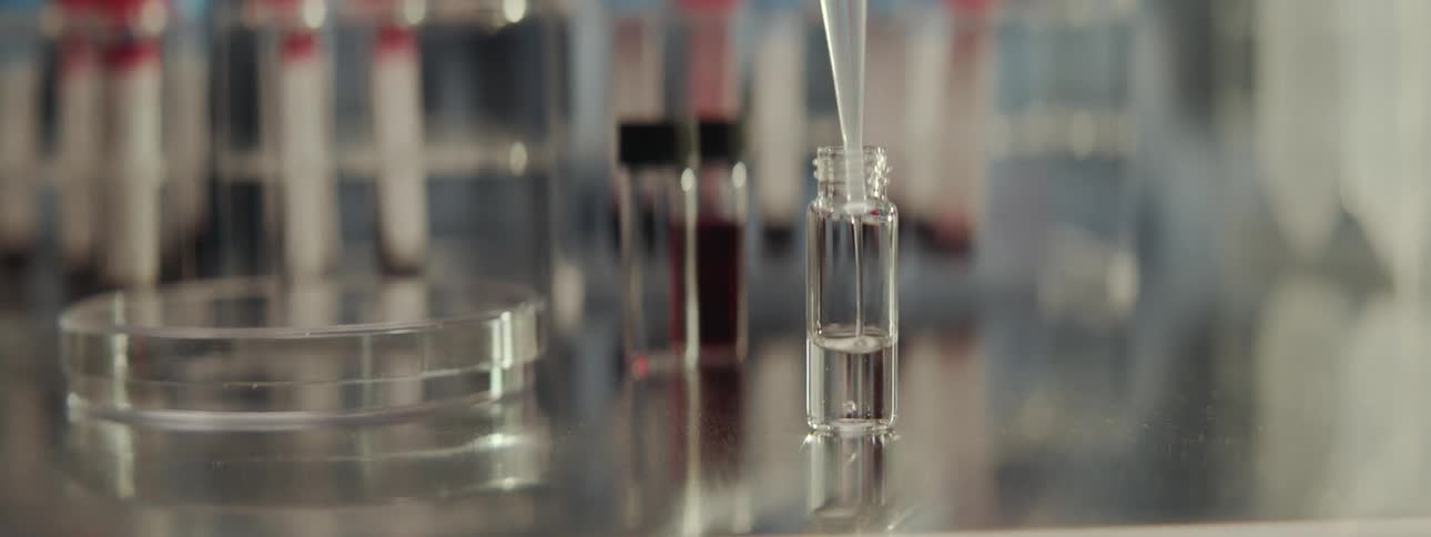 Slow motion close-up of a pipette carefully dispensing a clear liquid into a glass vial in a scientific laboratory