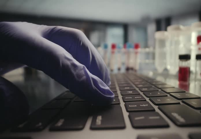 A scientist in blue gloves types on a laptop keyboard in a laboratory with test tubes in the background