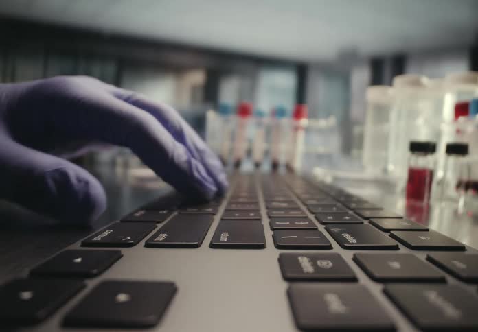 Close-up of a gloved hand typing on a keyboard in a scientific laboratory with test tubes in the background