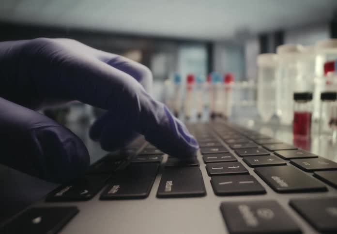 A scientist in blue latex gloves types on a laptop keyboard in a research laboratory with test tubes in the background
