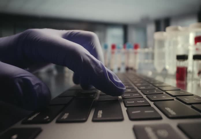 A close up of a researcher's gloved hands typing on a laptop keyboard inside of a laboratory with vials in the background