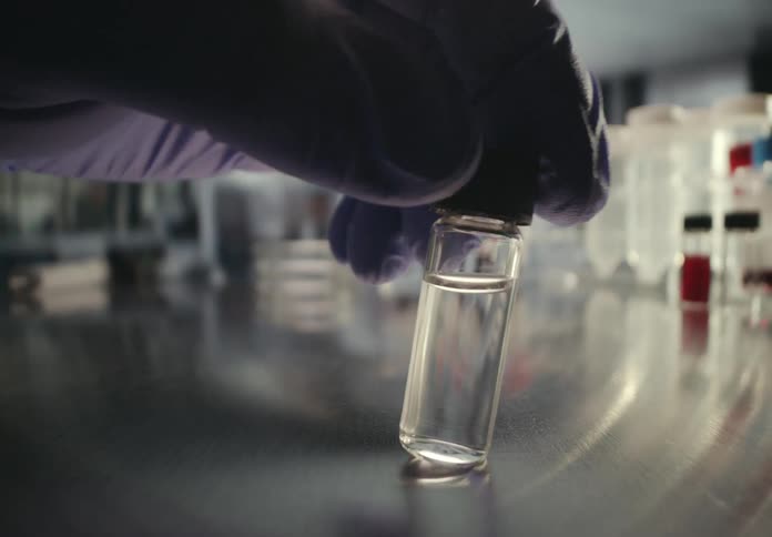 A scientist's gloved hand carefully examines a small vial of clear liquid on a steel counter in a research lab