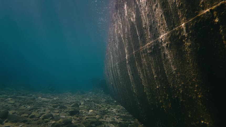 An underwater view shows the hull of a sunken shipwreck resting on the rocky seabed as sunlight filters through the water