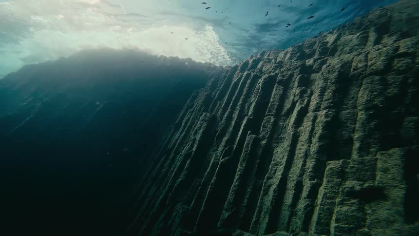 Sunlight illuminates a majestic underwater rock formation as waves ripple on the surface above