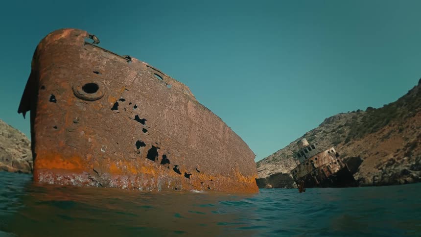 Water-level view of two massive, rusted shipwrecks slowly decaying in a secluded, rocky bay under a clear sky