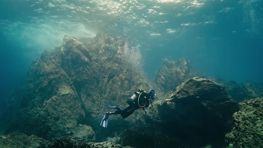 A scuba diver floats peacefully underwater near a rocky reef as bubbles rise to the sunlit surface in this looping cinemagraph