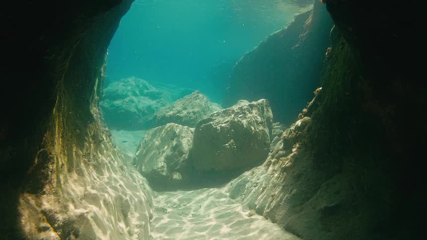 A serene underwater view from inside a rocky cave shows sunlight dancing on the sandy ocean floor