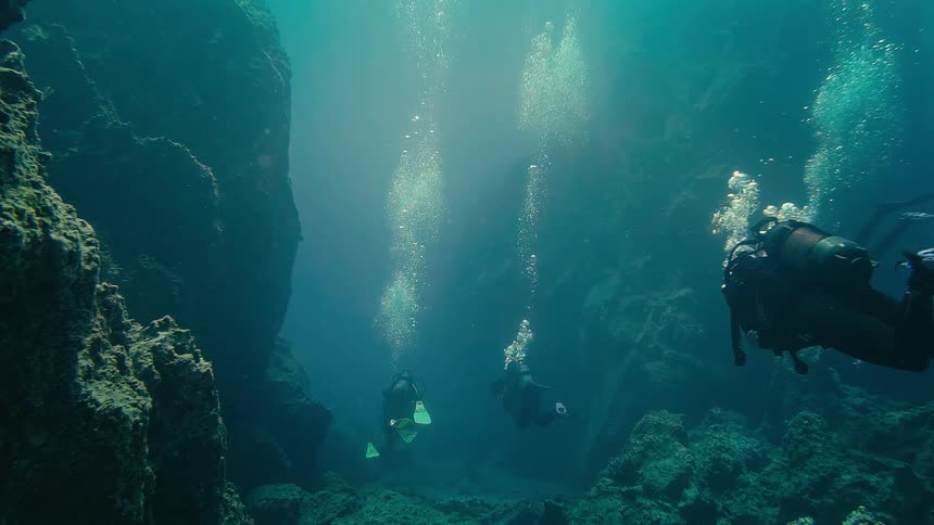 Three scuba divers float peacefully in a deep, dark underwater canyon as bubbles rise and sunlight shines from above