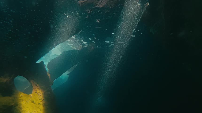 An underwater view from a dark grotto looks up at sunbeams filtering through floating ice and rising bubbles