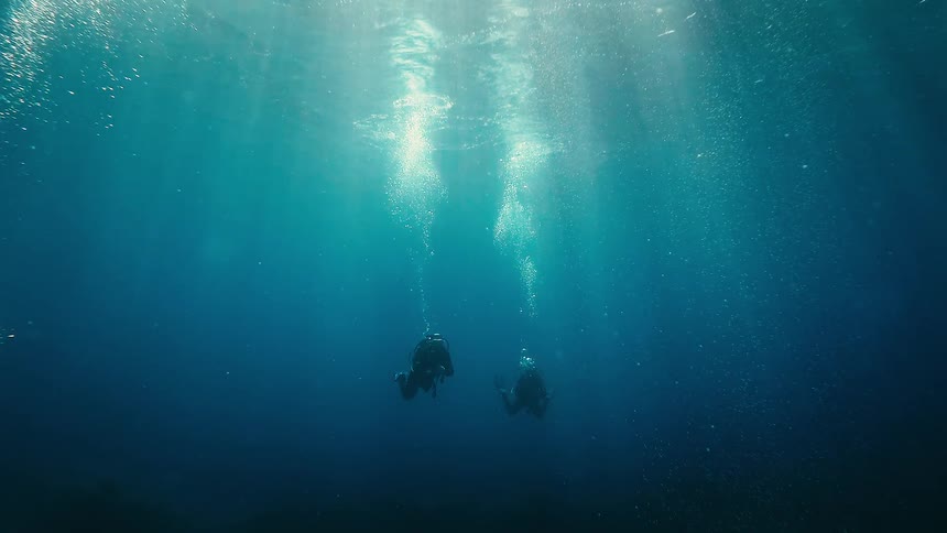 A serene underwater view shows two scuba divers floating in the deep blue ocean, illuminated by beautiful sun rays from above