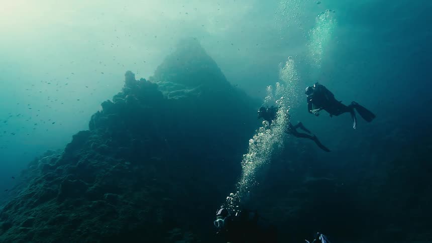 Three scuba divers explore a massive underwater rock formation as bubbles rise to the sunlit surface in a seamless loop
