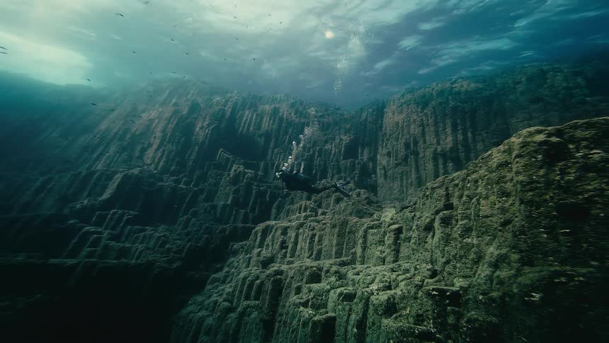 A scuba diver floats serenely while exploring the vast, algae-covered walls of a dramatic underwater canyon