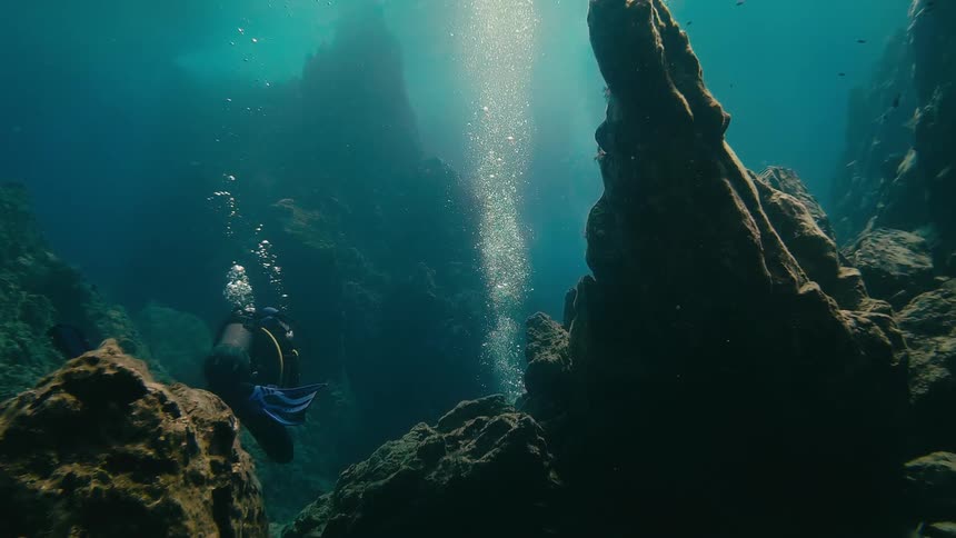 A scuba diver explores a deep underwater canyon as bubbles rise and sunlight beams down from the surface