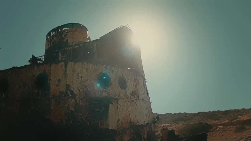 The bright sun backlights a rusted, decaying shipwreck beached on a desolate coast