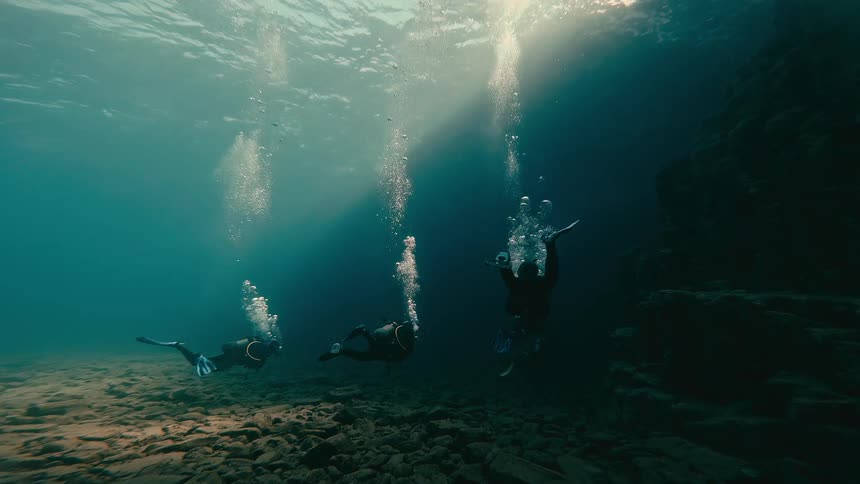 Three scuba divers explore the rocky ocean floor as bubbles rise towards the sunlit surface