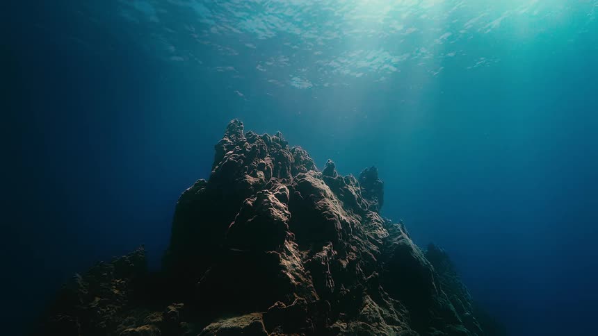 Sunlight streams down into the deep blue ocean, illuminating a massive underwater rock formation
