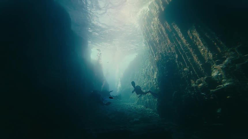 Scuba divers swim through a narrow underwater canyon as beautiful sunbeams shine down from the surface
