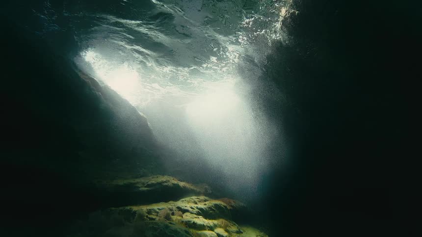 A cinematic view from inside an underwater cave shows ethereal light rays piercing the dark water from an opening at the surface